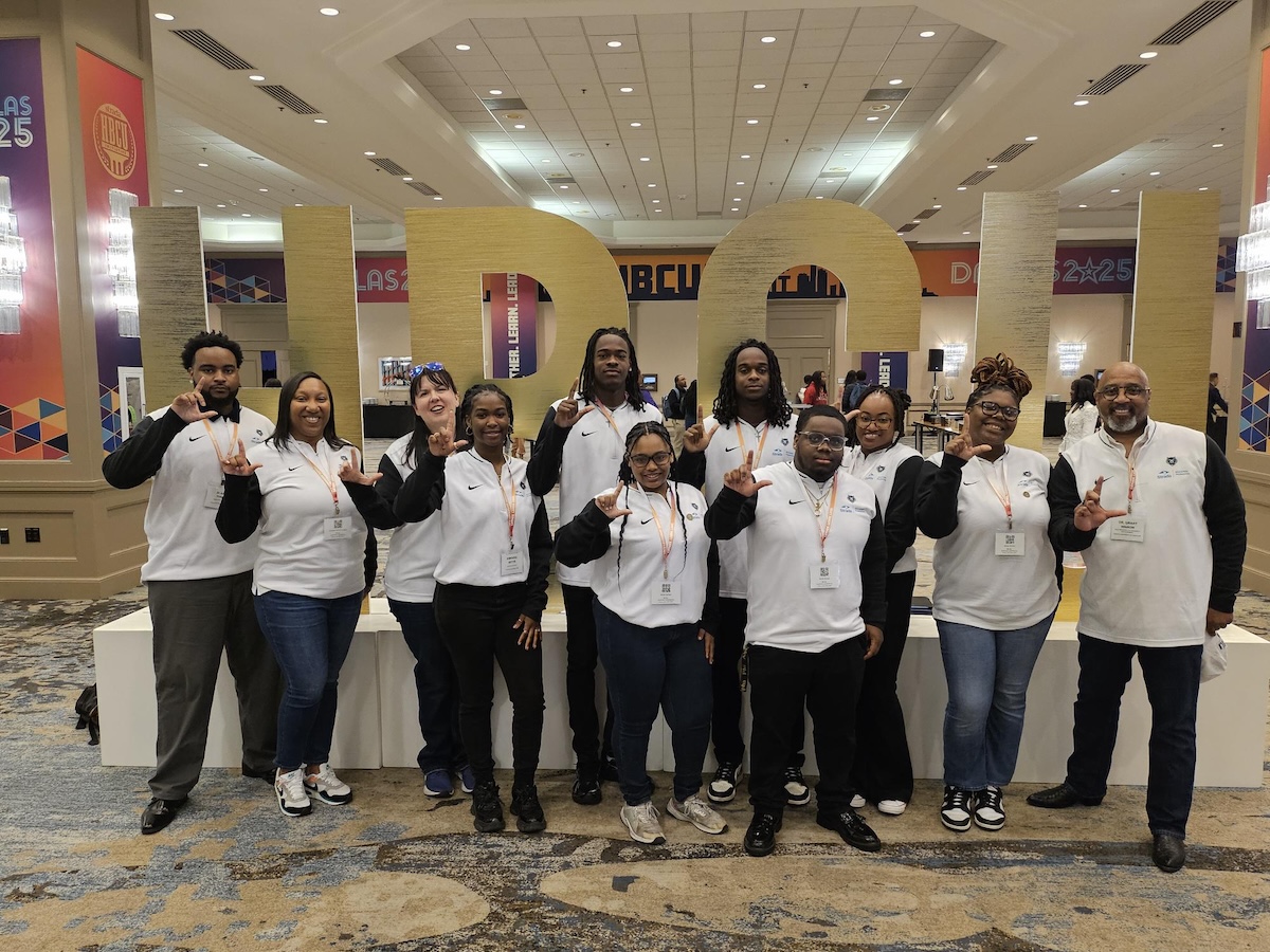 Group photo of Lincoln University Strada Scholars and program advisors standing in front of large gold 'HBCU' letters at a conference. All individuals are wearing matching white and black Strada shirts and making the 'L' hand sign for Lincoln